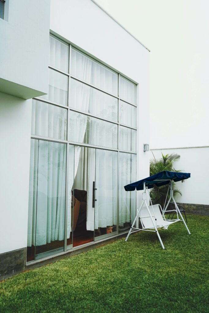 pexels-photo-16631165-16631165 Modern home exterior with large glass door and swing chair on a green lawn.